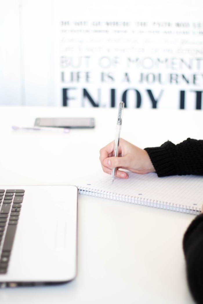 Close-up of a person's hand writing notes in an office setting with a laptop.